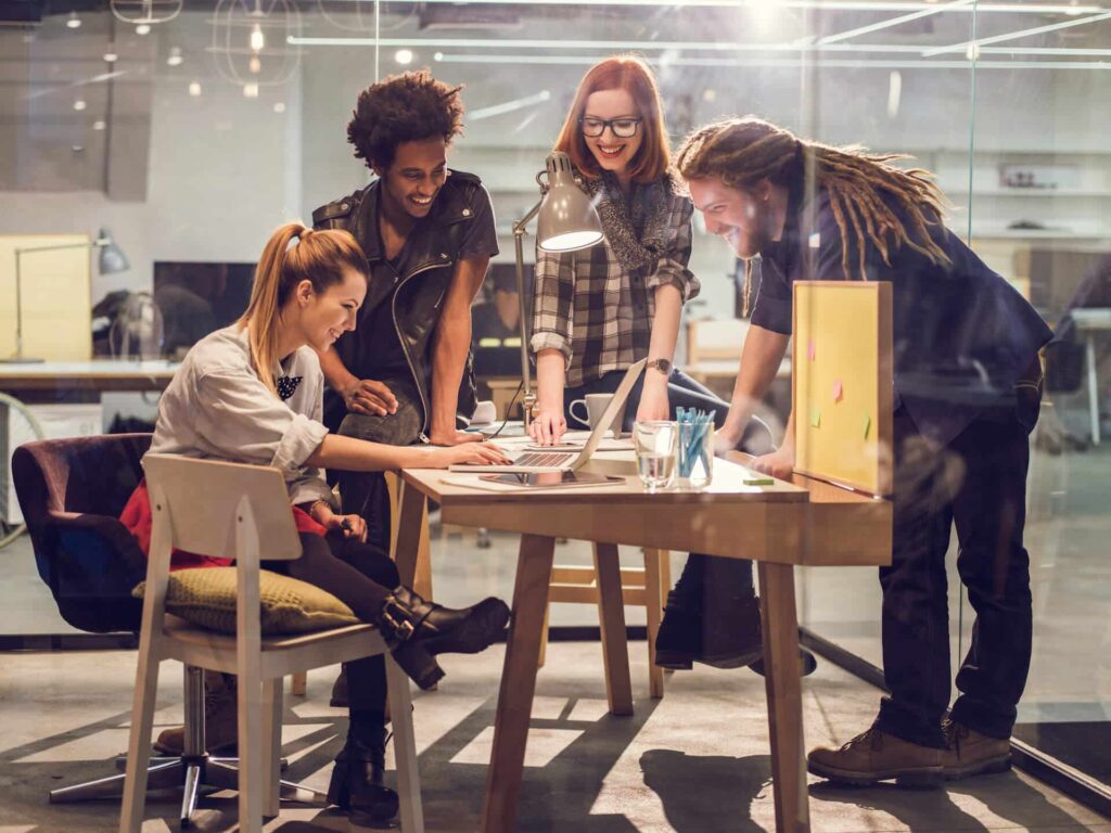 Four people collaborating around a laptop at a modern office table, looking engaged and smiling.