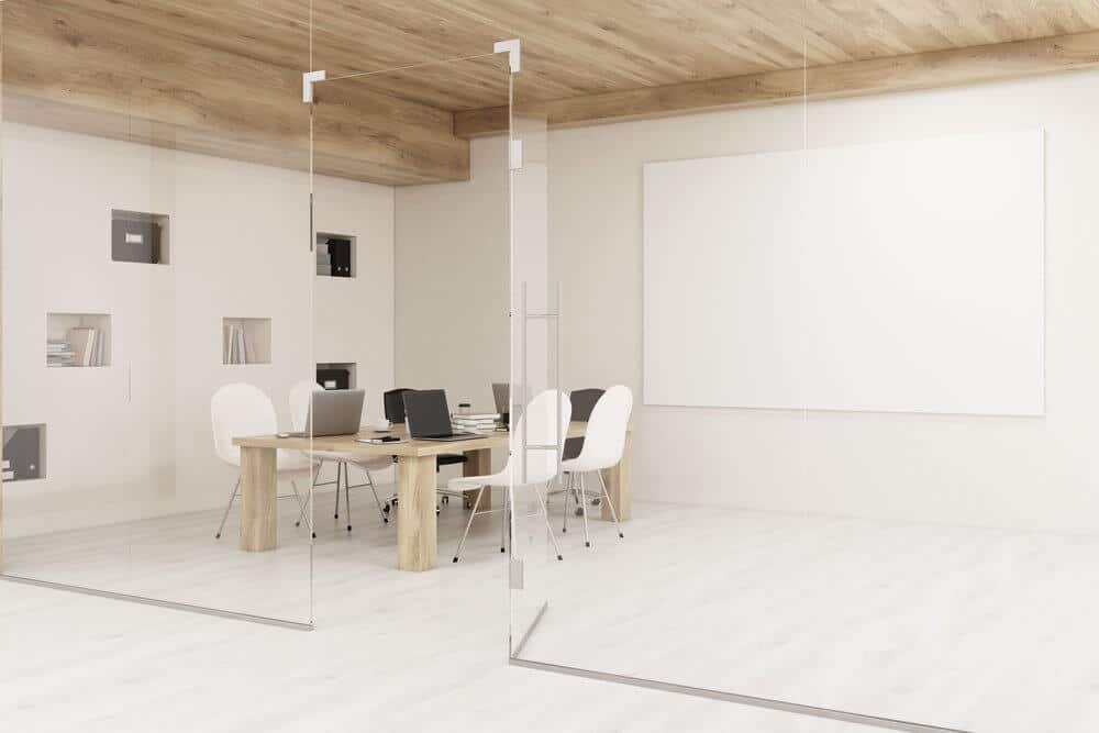 Minimalist office with a wooden table, white chairs, laptops, and large whiteboard, enclosed by glass walls.