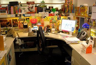 Colorful, cluttered office cubicle with computer screens, books, papers, and various decorations on walls and desk.