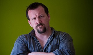 Person with a contemplative expression, arms crossed, wearing a blue shirt against a green background.