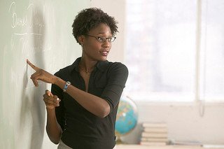 A woman stands at a blackboard pointing, with a globe and books in the background.