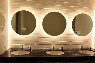 Three round mirrors above sinks on a dark countertop, set against a wall with white tiles.