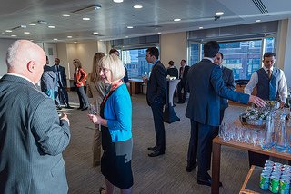 People mingling in a conference room with refreshments, wearing business attire, and engaged in conversation.