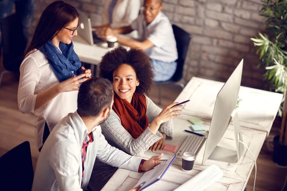 A group of people smiling and collaborating at a desk with a computer and papers in a bright office setting.
