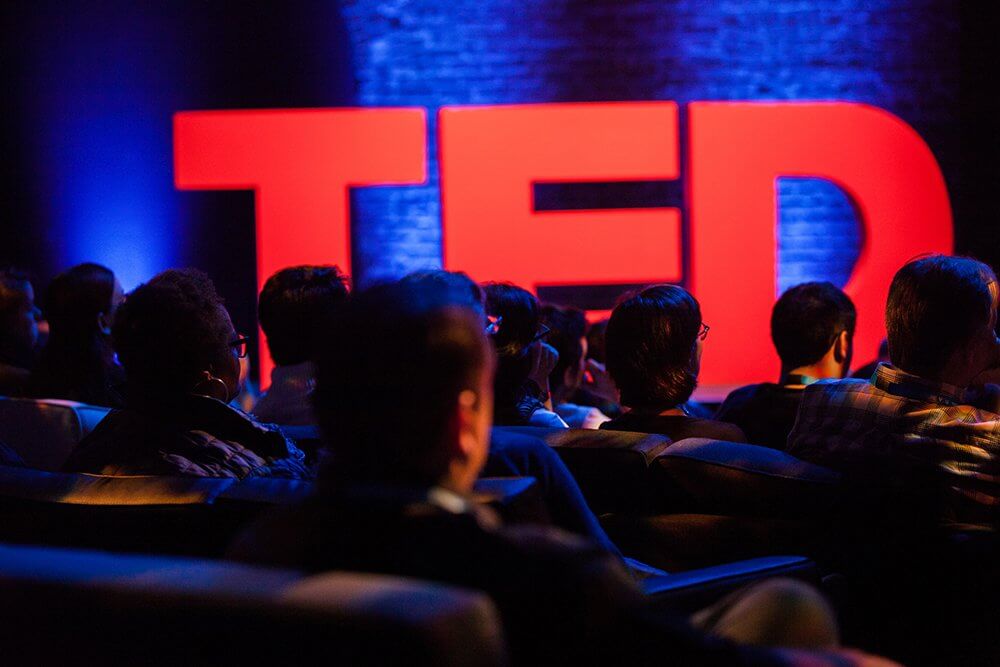 Audience in a dimly lit room watches a presentation with large red "TED" letters in the background.