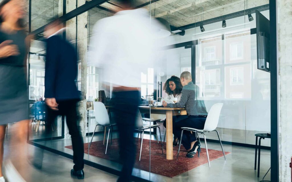 Blurry motion of people walking in a modern office with two people sitting at a table inside a glass-walled room.