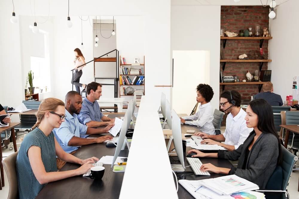 Open office with diverse people working at computers and collaborating. Brick wall and bookshelves in the background.