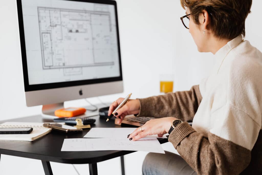A person working at a desk with a computer displaying architectural plans, holding a pen and paper.