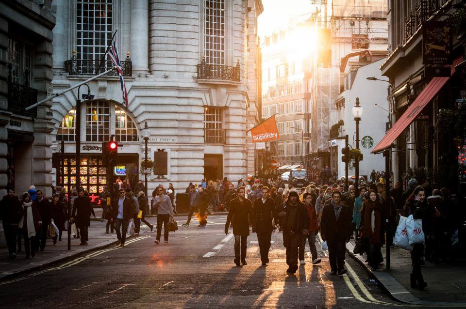 A bustling city street at sunset with people walking and historic buildings in the background.