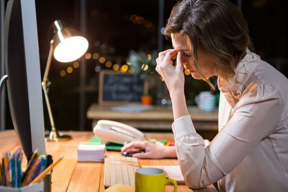 A woman sits at a desk, rubbing her forehead, looking stressed, with a computer and lamp illuminating the scene.