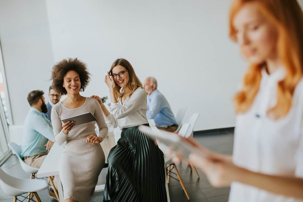 People in a bright office, some seated and some standing, engaged in conversation and using electronic devices.