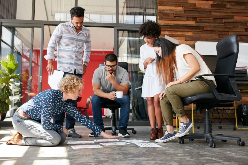 Five people discussing documents on the floor in a modern office setting with a large wooden wall.