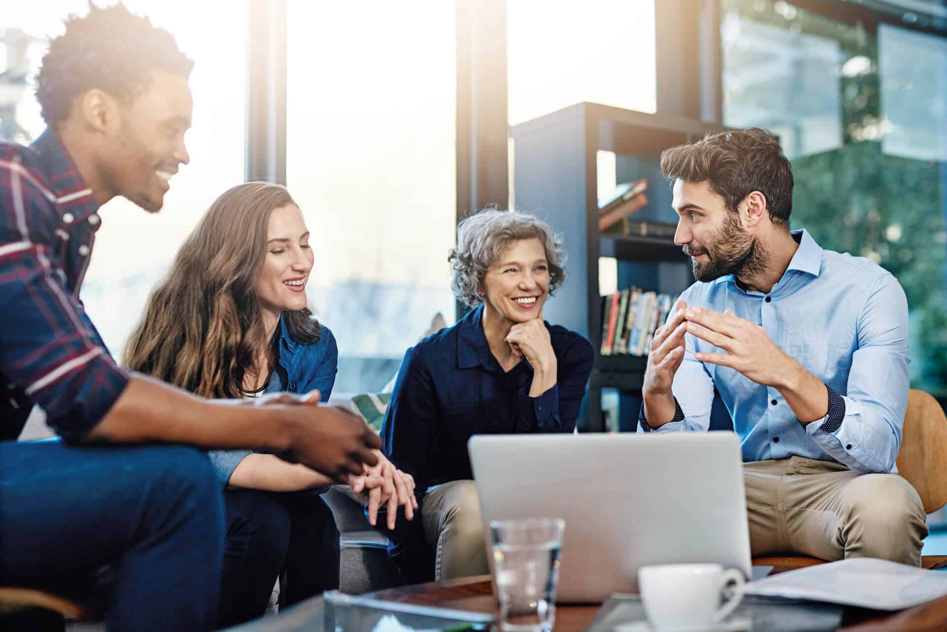 Diverse group of four people smiling and discussing around a laptop in a modern office setting.