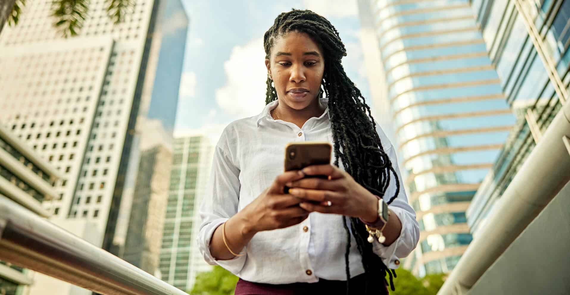 Woman with long braids using a smartphone outside, with tall modern buildings in the background.