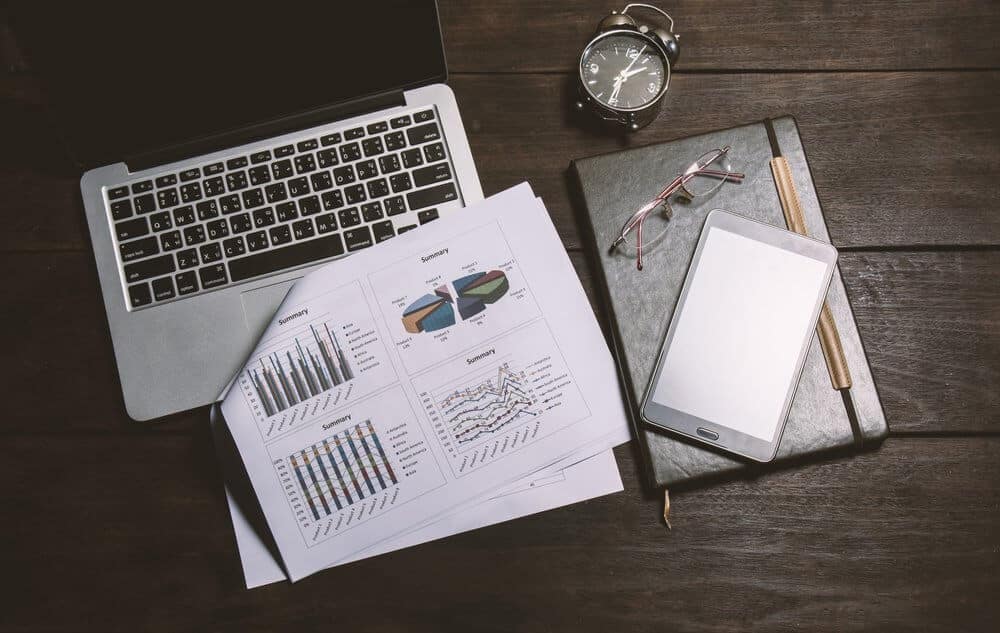 Laptop, financial charts, clock, tablet, and glasses on a wooden desk, suggesting a work or study setting.