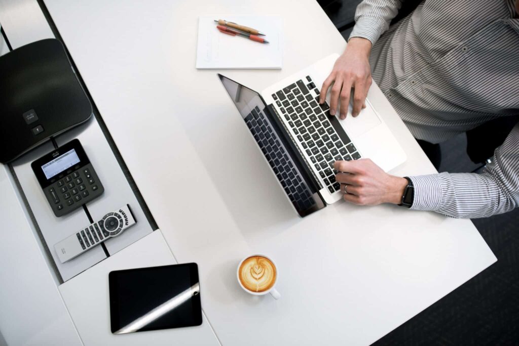Person working on a laptop at a desk with a notepad, tablet, phone, and a cup of coffee nearby.