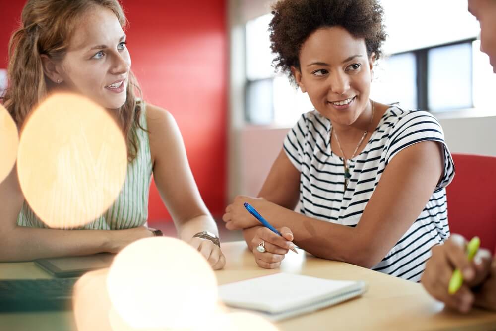Two people sitting at a table, smiling and talking, with notebooks and pens in a well-lit room.