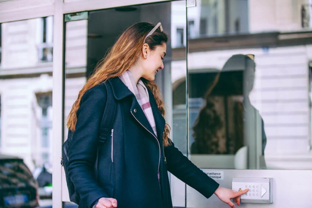 A woman with sunglasses on her head enters a security code on an outdoor keypad by a glass door.