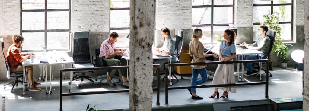 Open office with six people working at desks, two standing and talking, large windows, and indoor plants.