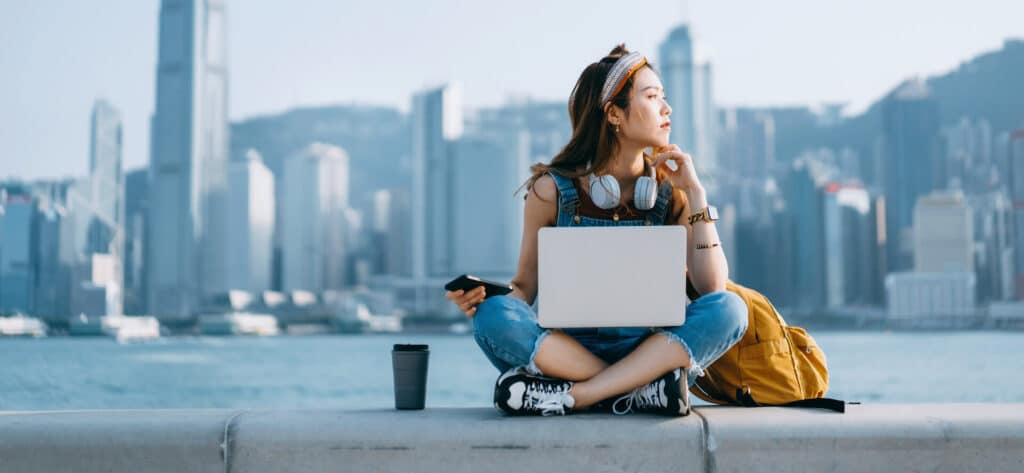 Woman sitting cross-legged by water with a laptop and headphones, city skyline in the background, looking sideways.