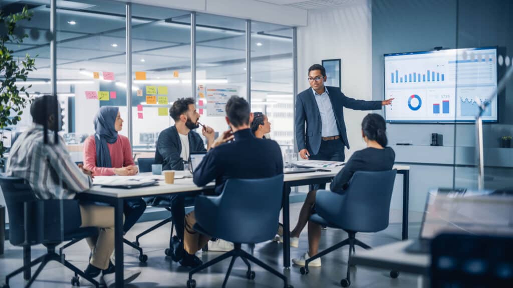 A person presents data on a screen to a diverse group seated around a conference table in a modern office.