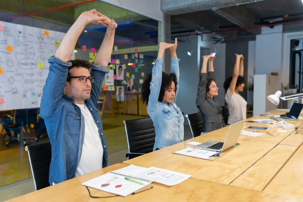 Four people seated at a conference table stretch with arms overhead in an office with diagrams and notes on the wall.