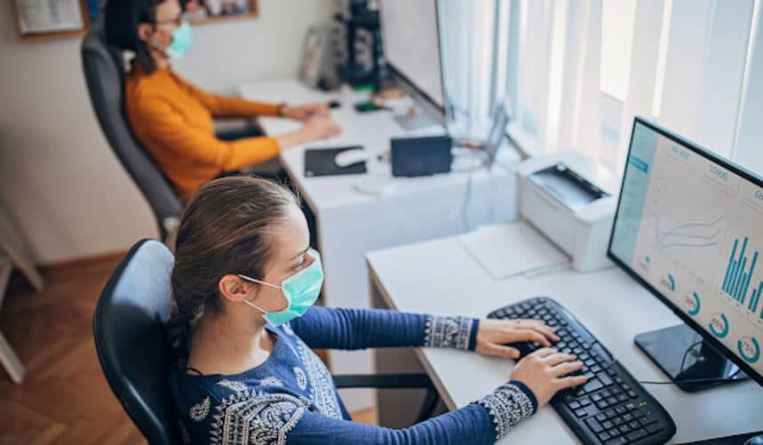 Two people wearing masks work at separate desks with computers in an office. One monitors graphs on a screen.