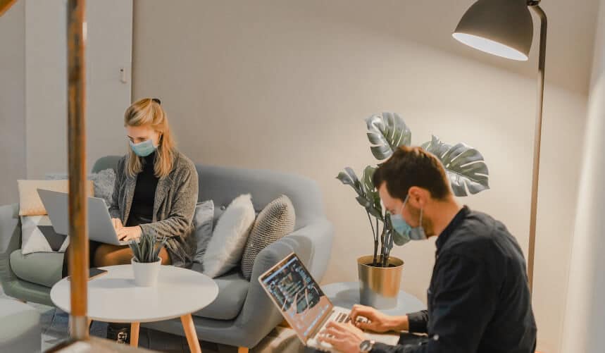 Two people wearing masks work on laptops in a cozy room with a sofa, plant, and floor lamp.