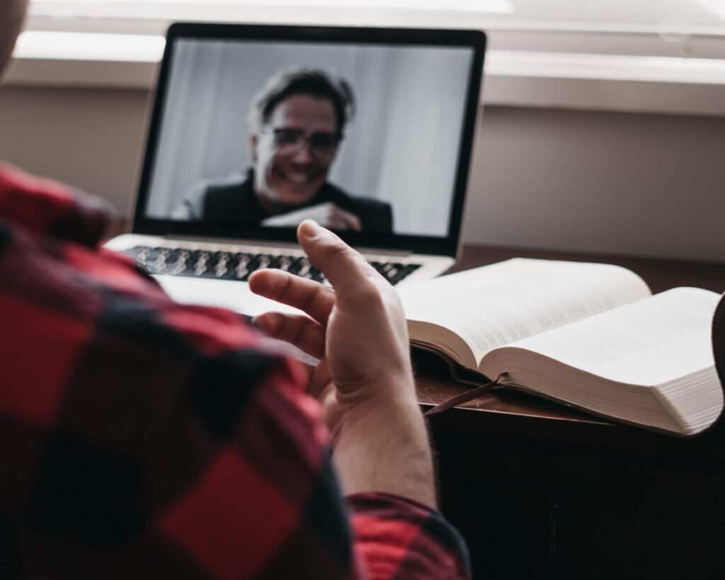 Person in a plaid shirt on a video call, with an open book nearby on a wooden desk using zoom integration
