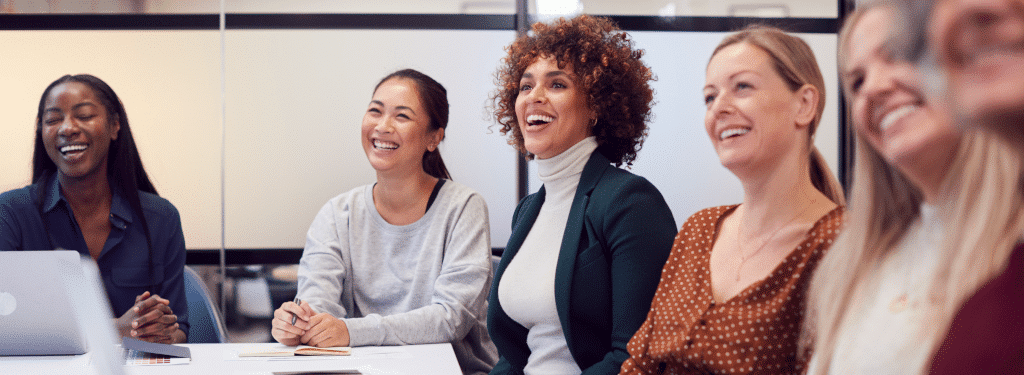 A diverse group of people seated at a conference table, smiling and attentively listening in a bright meeting room.