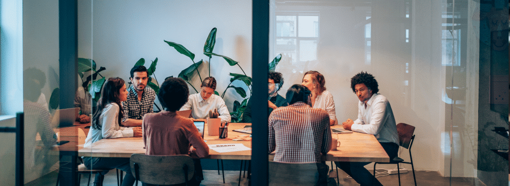 A group of people having a meeting in a modern conference room with large plants in the background.