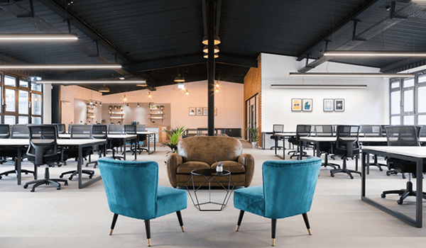 Modern open-plan office with teal chairs, a brown sofa, desks, and ceiling lights.