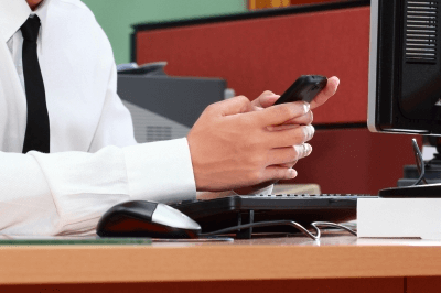 Person in a white shirt text messaging on a phone at an office desk with a computer.
