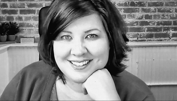 Woman smiling, resting her chin on her hand, sitting in front of a brick wall background. Black and white image.