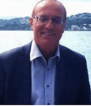 A man in glasses and a suit smiles, standing in front of a body of water with hills and houses in the background.