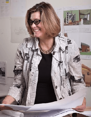 Smiling woman in glasses holds architectural plans, with project drawings on the wall behind her.