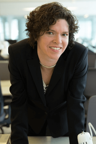 Person in a black suit smiling and leaning on a desk in a bright office setting.