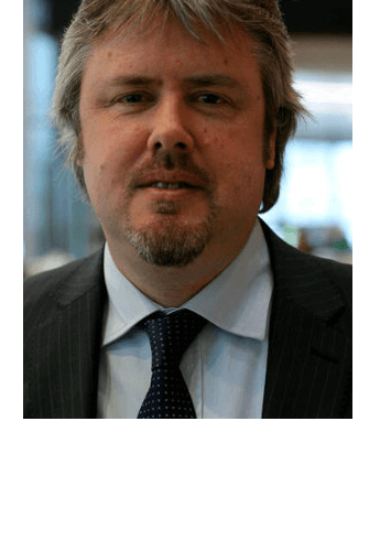 Bearded man in a suit and tie, standing indoors, with blurred background.