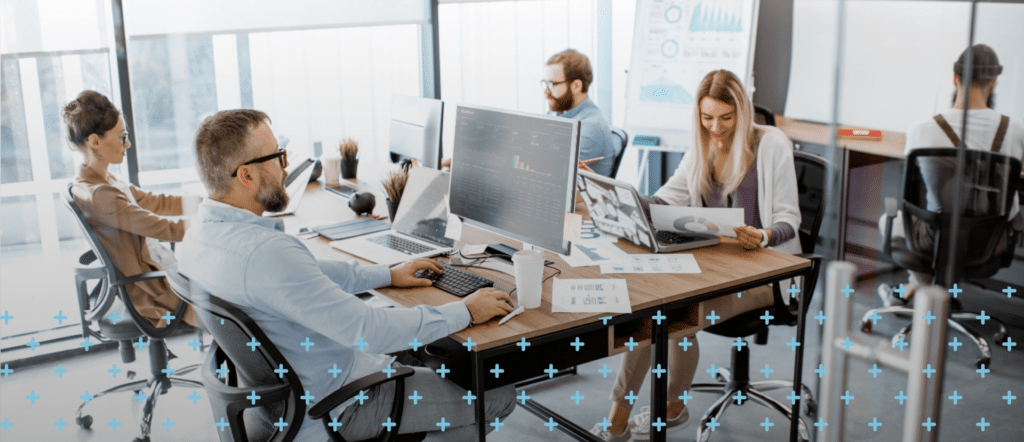 Open office with four people working at desks, one on a computer, others reading documents, and person in background.