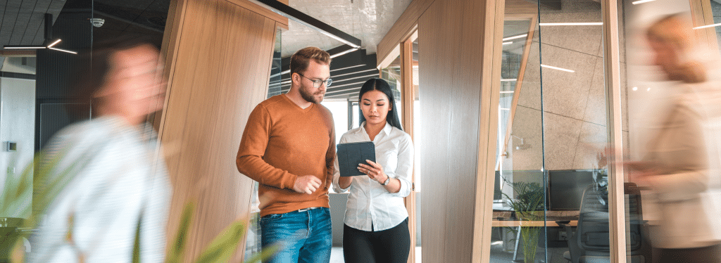 Two coworkers walking in an office, focused on a tablet, with blurred colleagues passing by.